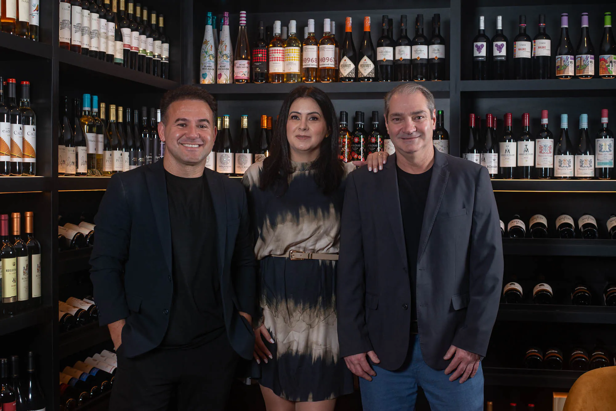 Rafael Moura, Glenda Moya e Maurício Wingert, sócios da Ello Wines, lado a lado diante de uma parede de vinhos.